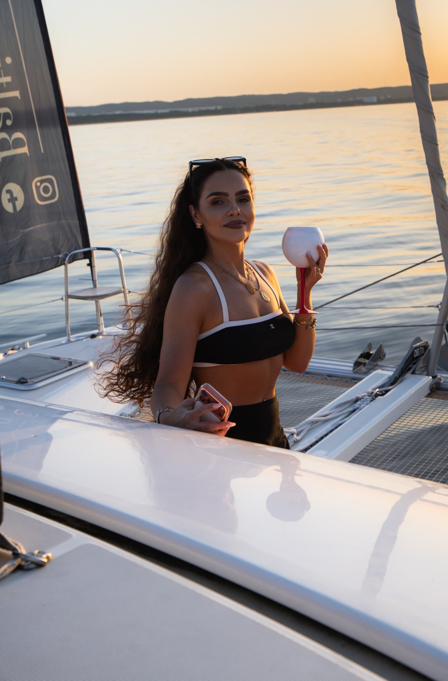 Kobieta w czarno-białym bikini trzyma kieliszek wina na tle morza o zachodzie słońca, elegancka poza na pokładzie katamaranu.