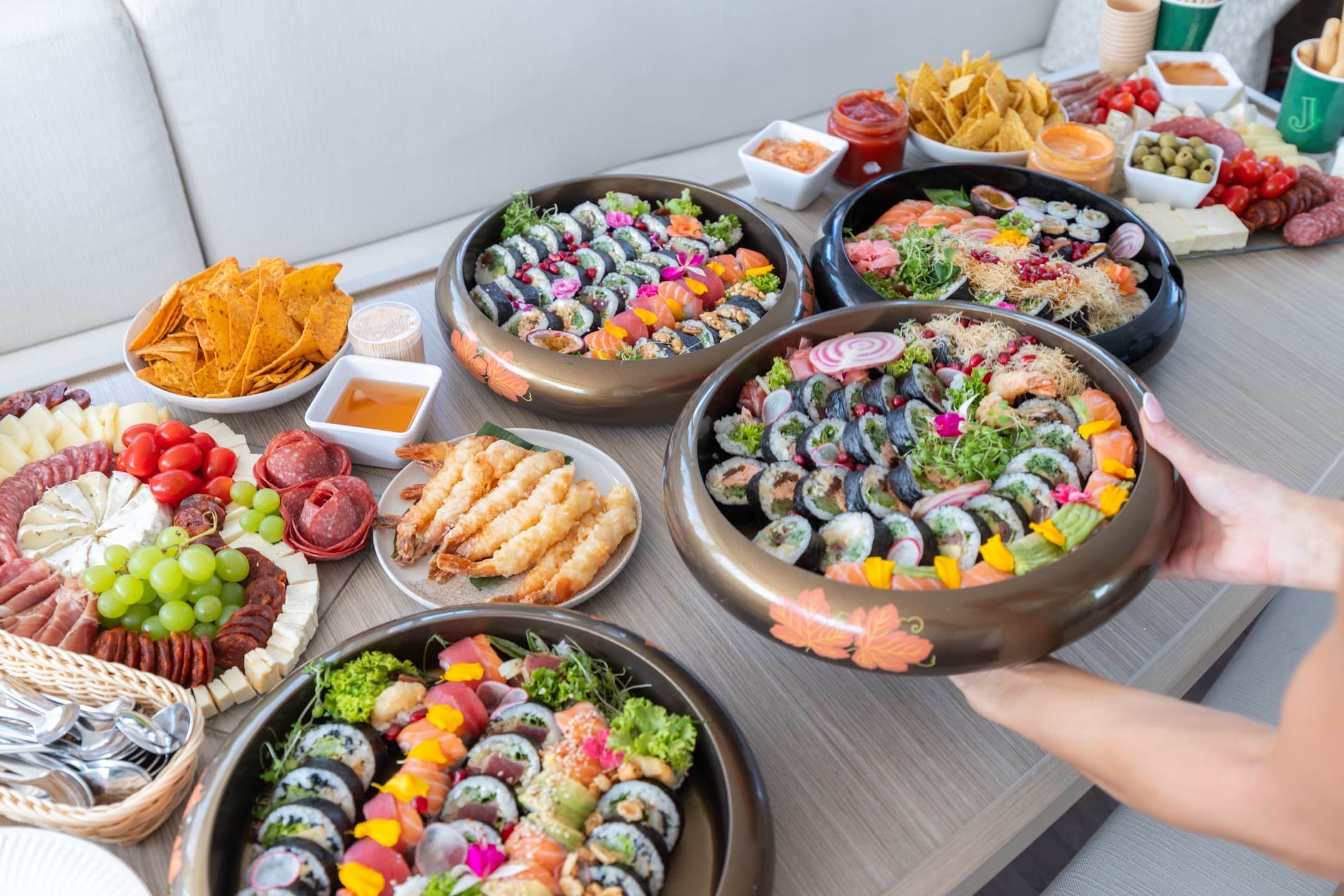 Rozłożone na stole tace sushi, deska serów i przekąski na pokładzie katamaranu, bogata kompozycja cateringowa w naturalnym świetle.