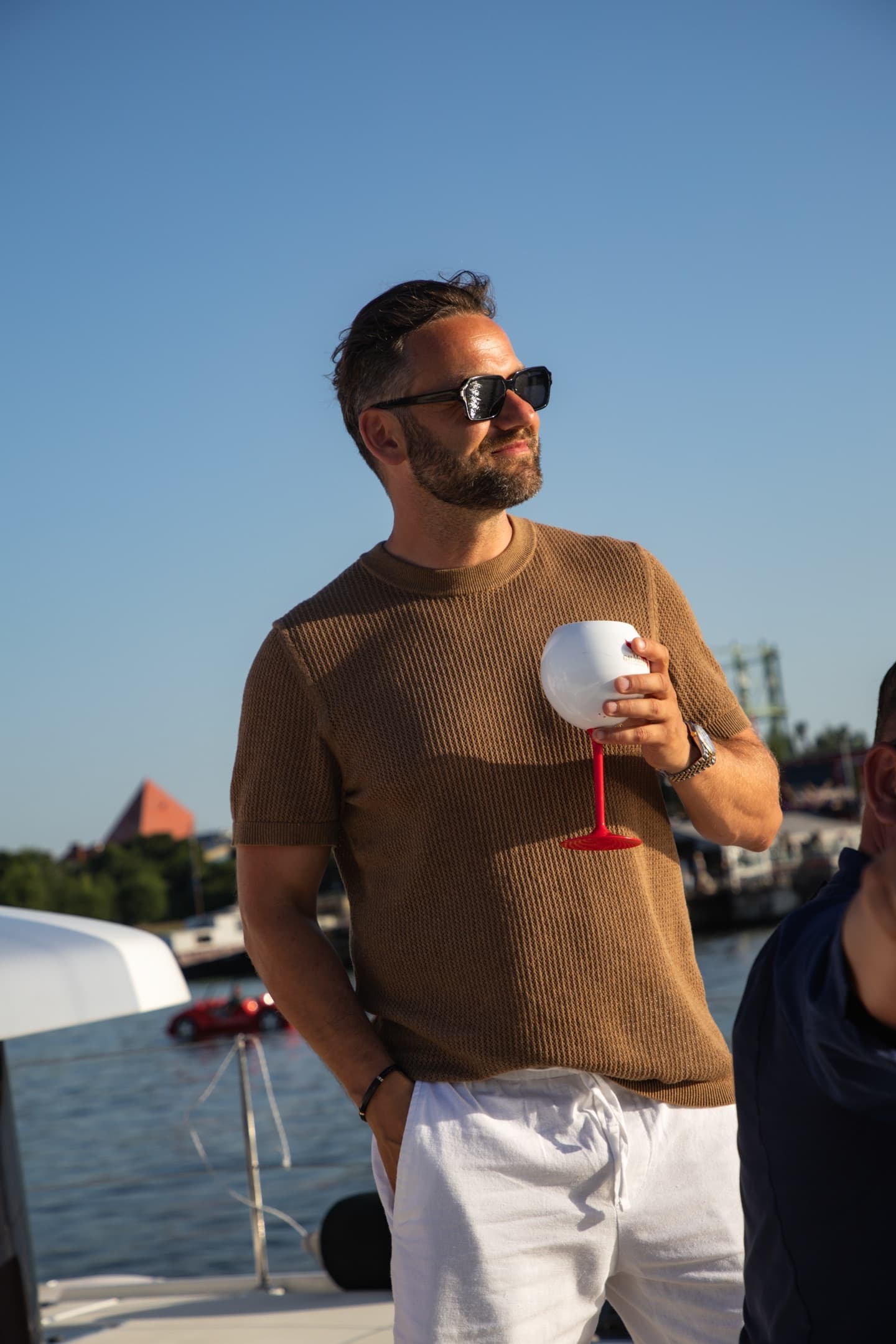 Elegancki mężczyzna w brązowym swetrze ze szklanką na czerwonej nóżce, spogląda w bok w ostrym słońcu, portowe tło.
