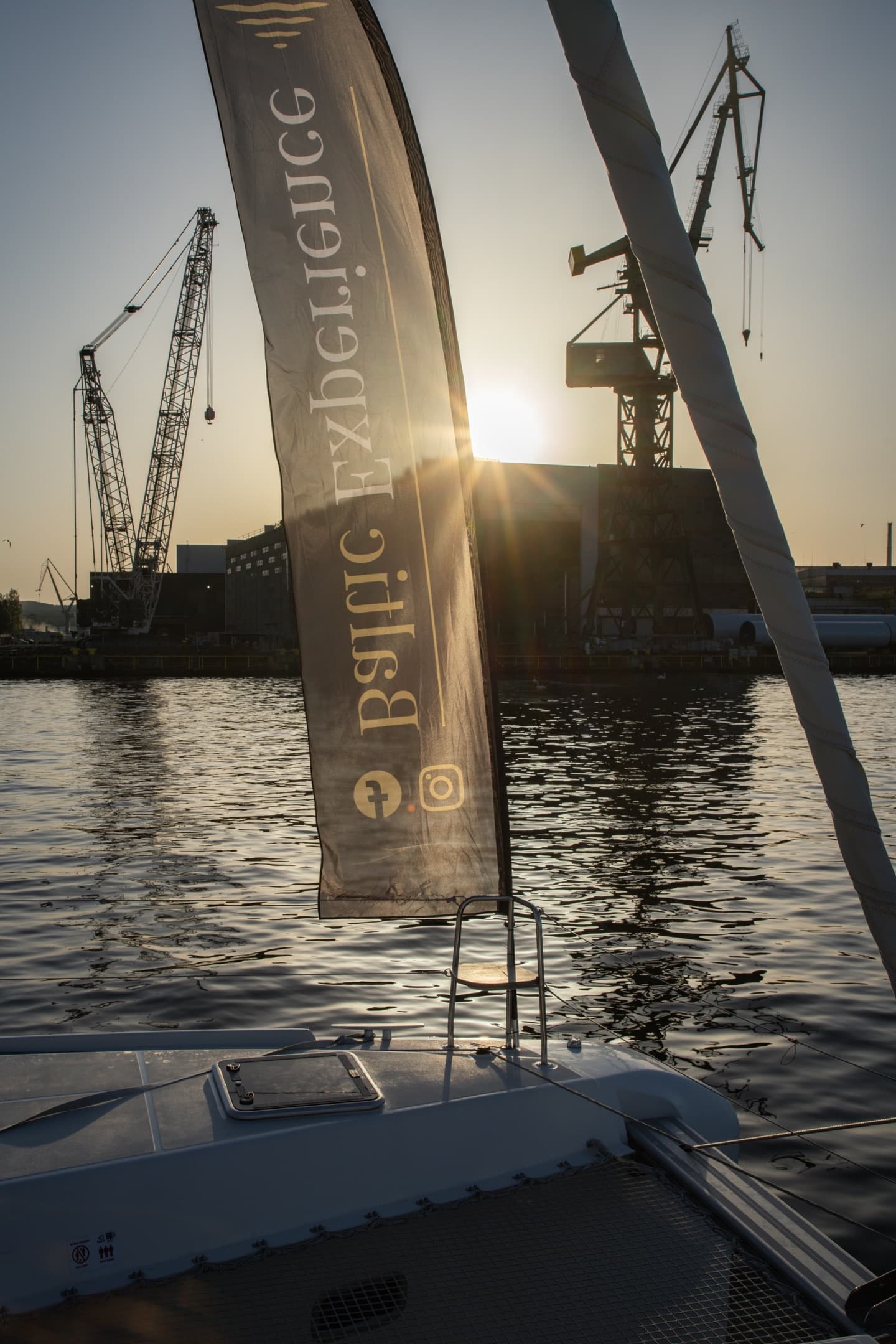 Flaga Baltic Experience na dziobie katamaranu na tle stoczniowych dźwigów i słońca przebijającego przez chmury, mocny branding.