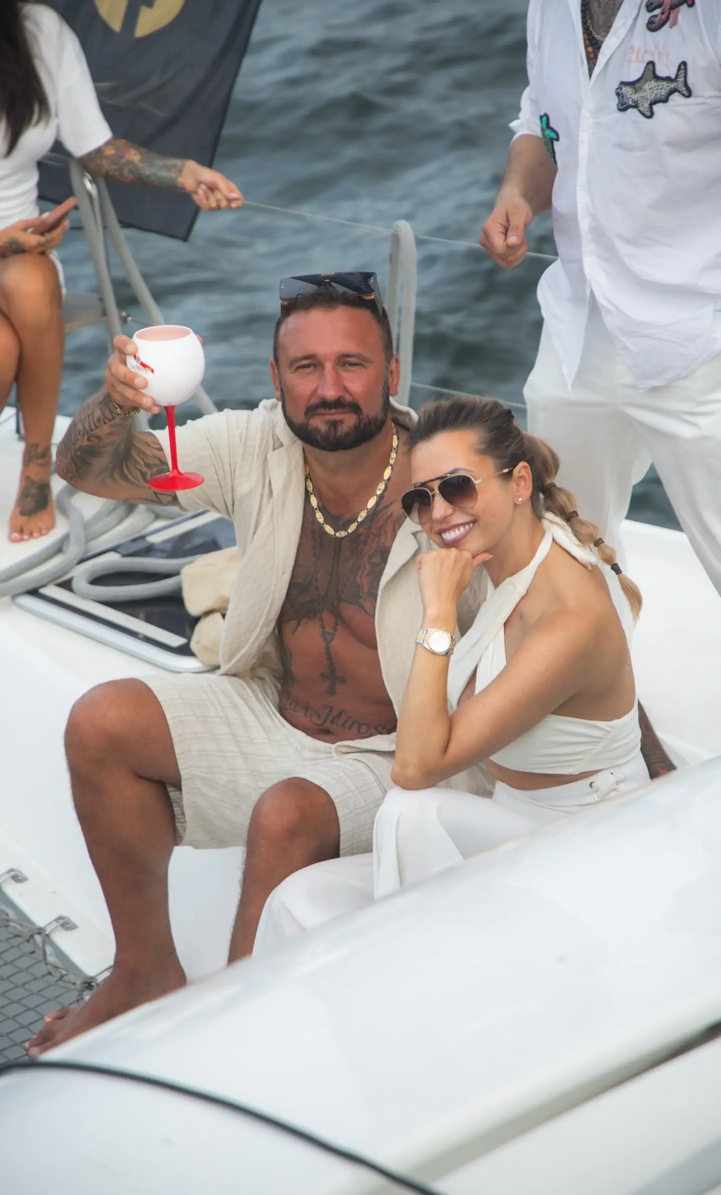 Para w eleganckich białych i beżowych strojach przytula się na pokładzie jachtu, on z kieliszkiem, oboje uśmiechnięci.