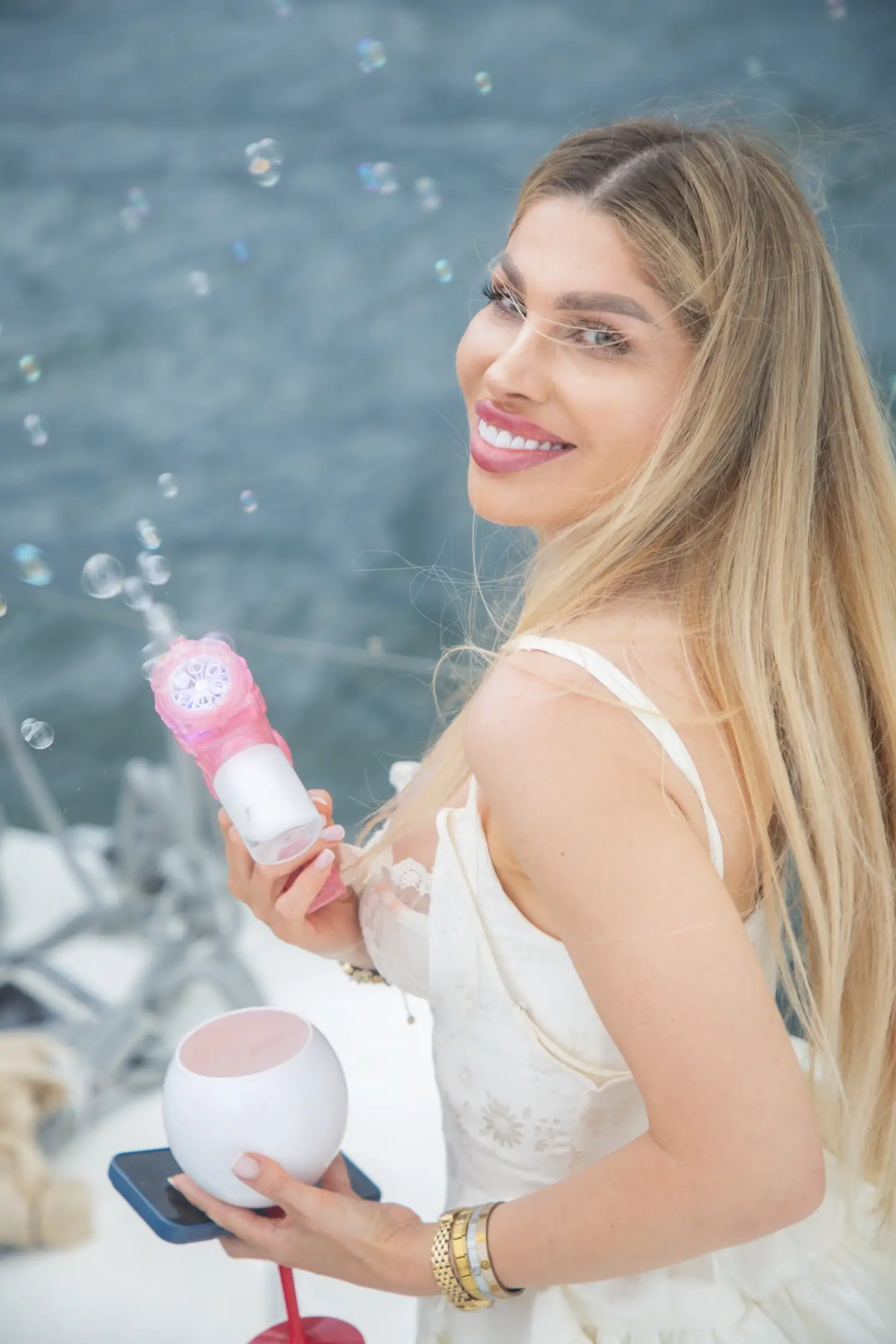 Blondynka w bieli z różową dmuchawą baniek, szeroki uśmiech, portretowe zbliżenie pełne radości.