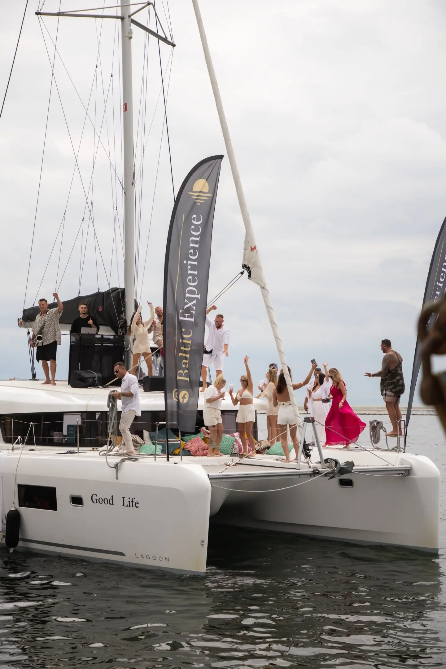 Szerokie ujęcie imprezy na katamaranie Good Life z flagą Baltic Experience, tańczący goście w bieli i kobieta w czerwonej sukience, DJ i saksofonista na pokładzie.