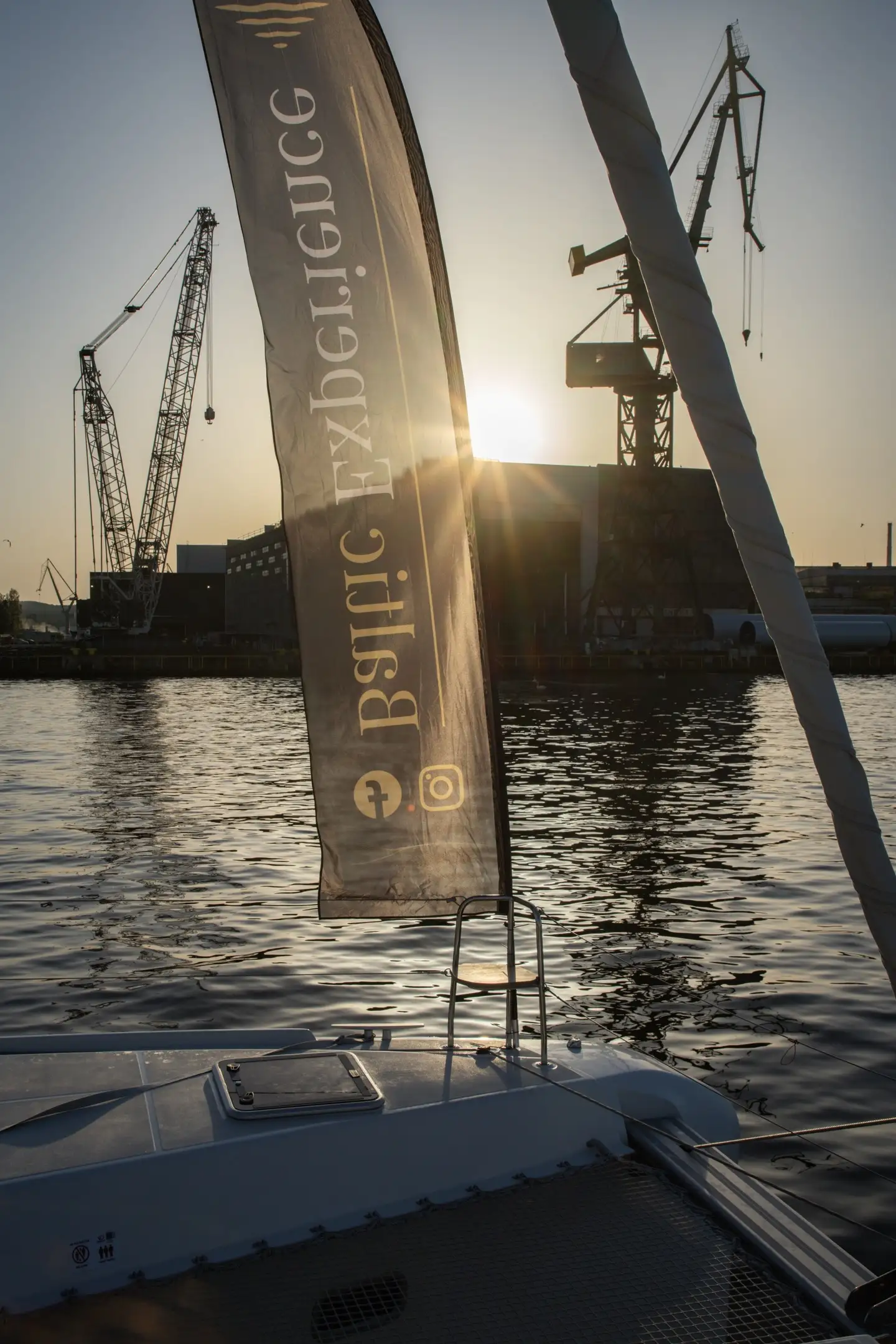 Flaga Baltic Experience na dziobie katamaranu na tle stoczniowych dźwigów i słońca przebijającego przez chmury, mocny branding.